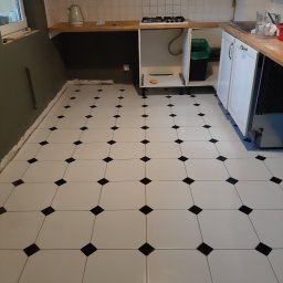 TK Format - Biało-czarna podłoga z płytek ułożona w kwadraty w kuchni w trakcie prac wykończeniowych, widoczne niedokończone fugi przy ścianie, fragment szafek kuchennych i sprzętów.