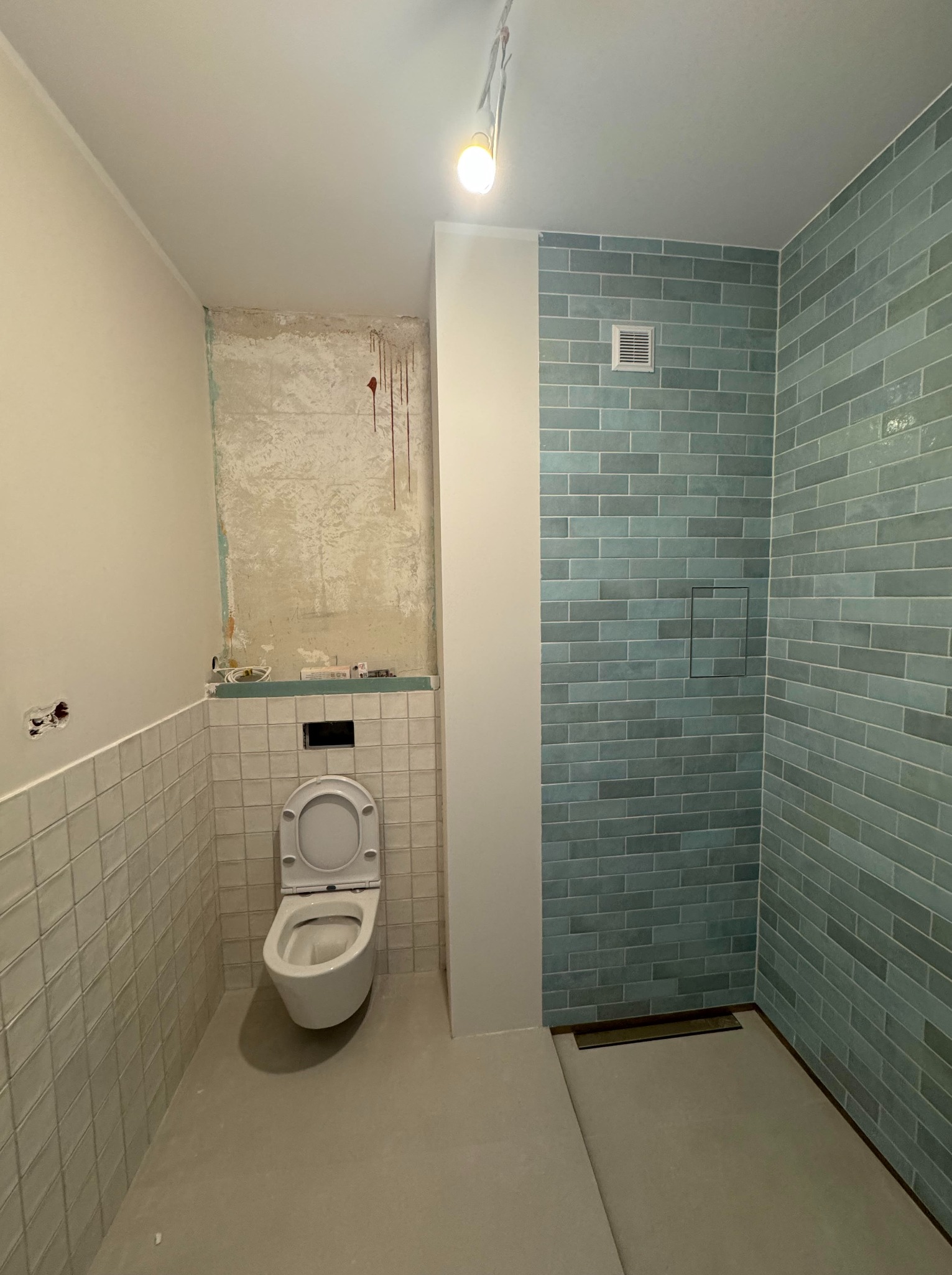 Remont łazienki w trakcie: częściowo wykończone ściany z płytek w odcieniach bieli i turkusu, otwarta toaleta, surowa ściana z instalacją.
