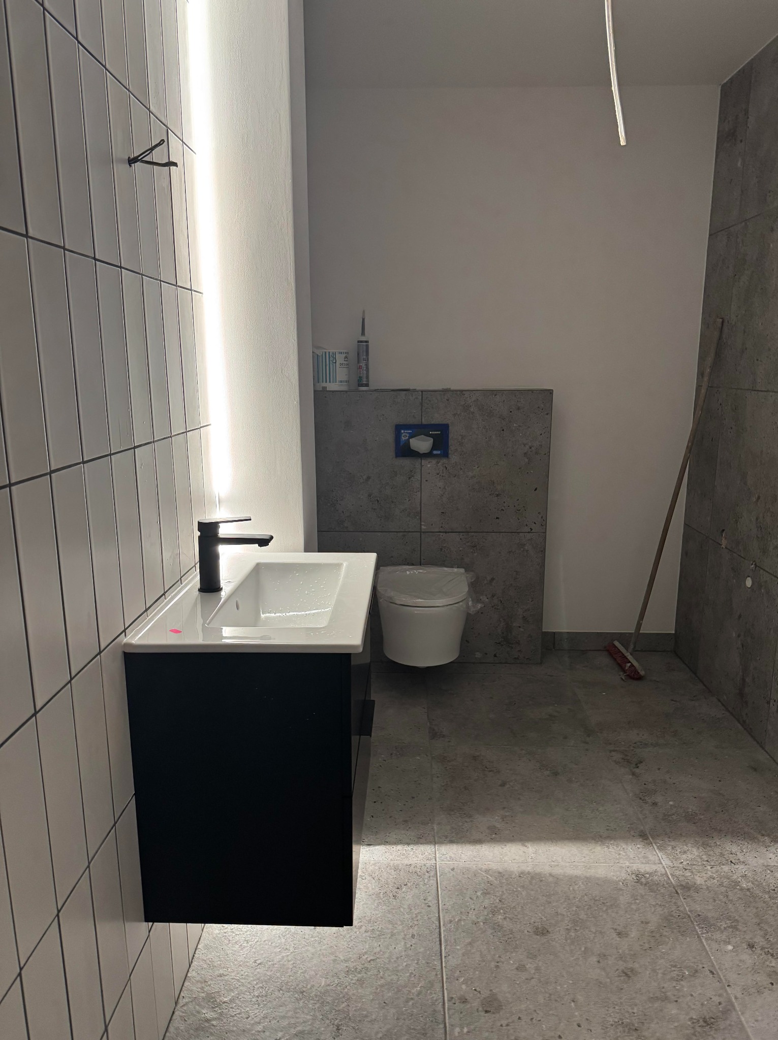 Nowoczesna łazienka w trakcie wykańczania: białe płytki, czarna szafka z umywalką, podwieszana toaleta i szare płytki imitujące beton.