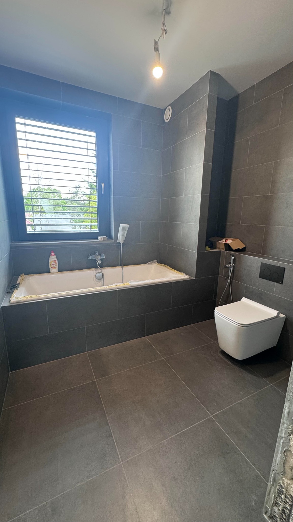 Wykończona łazienka w szarości. Nowoczesna toaleta, wanna zabezpieczona folią, okno z żaluzjami. Minimalistyczny design z ciemnymi płytkami i wiszącą lampą.