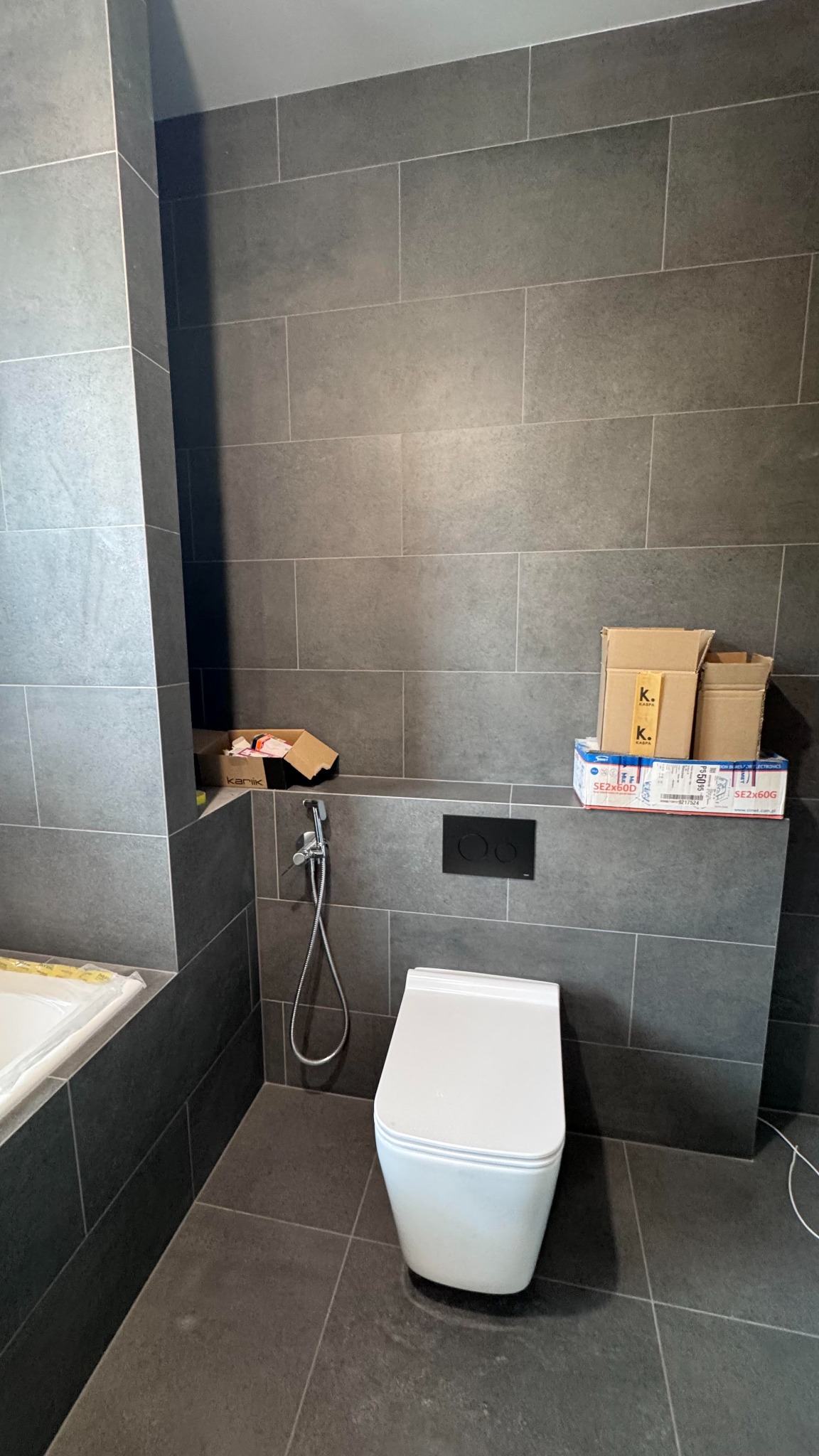 Nowoczesna łazienka z szarymi płytkami, wiszącą toaletą i bidetką. Na półce pudełka i opakowania po akcesoriach. Minimalistyczny styl, stonowana kolorystyka.