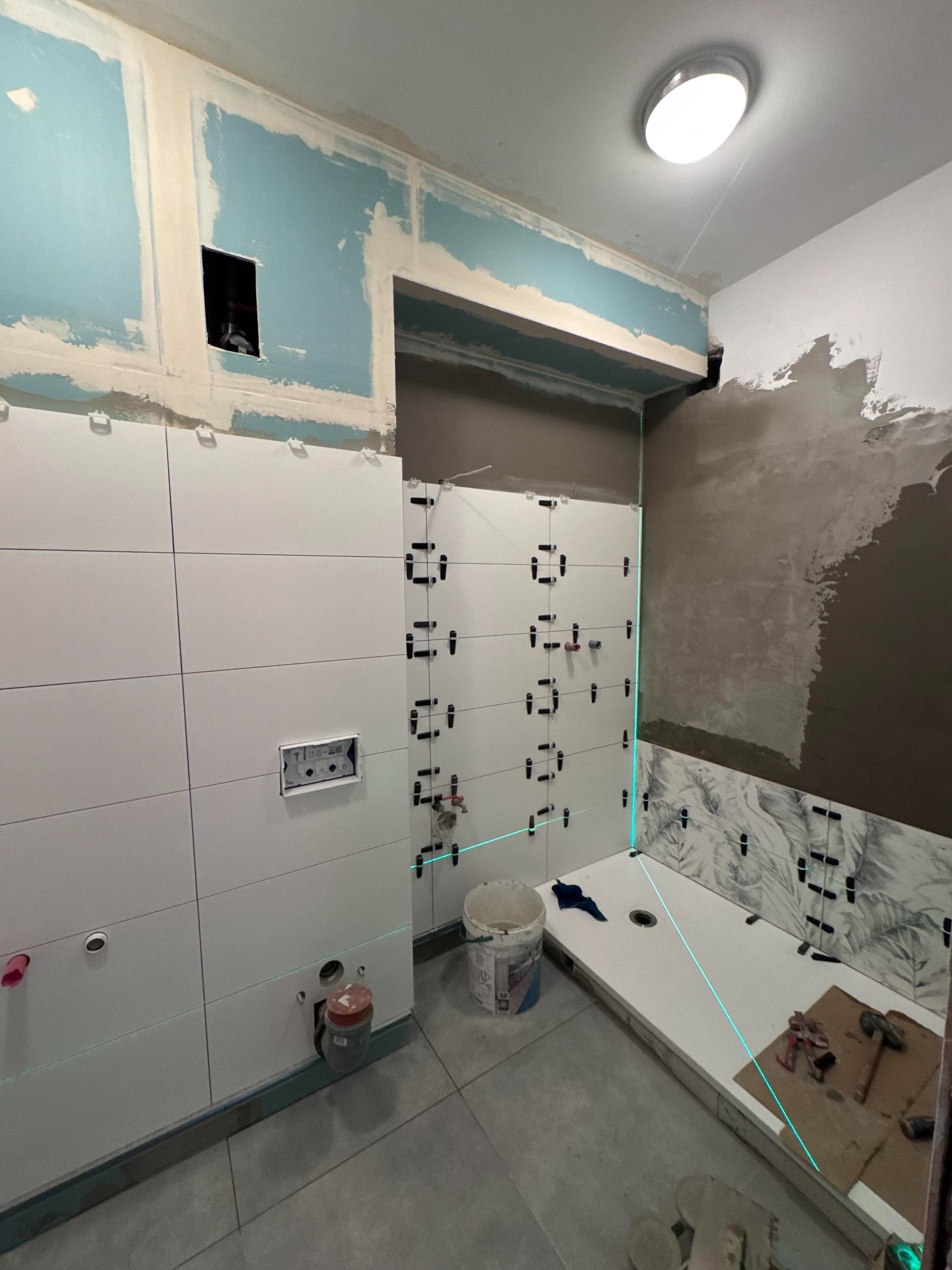 Białe płytki układane w łazience, z użyciem poziomicy laserowej i klipsów poziomujących. Częściowo wykończone ściany, widoczne narzędzia i materiały budowlane.