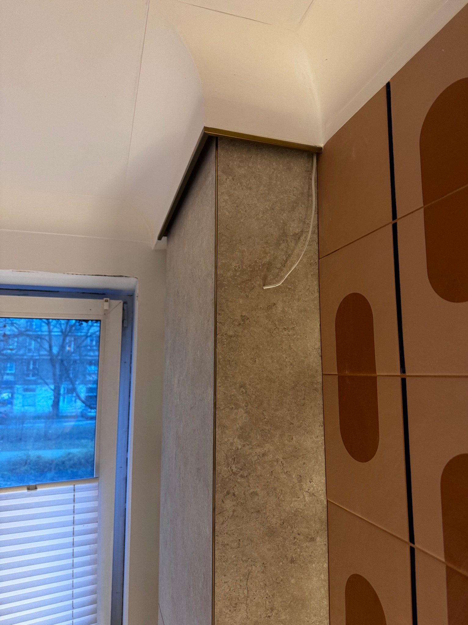 Wykończony narożnik ściany z płytami imitującymi kamień, z metalową listwą. Obok ściana z dekoracyjnymi panelami w odcieniach brązu. W tle okno z żaluzjami.