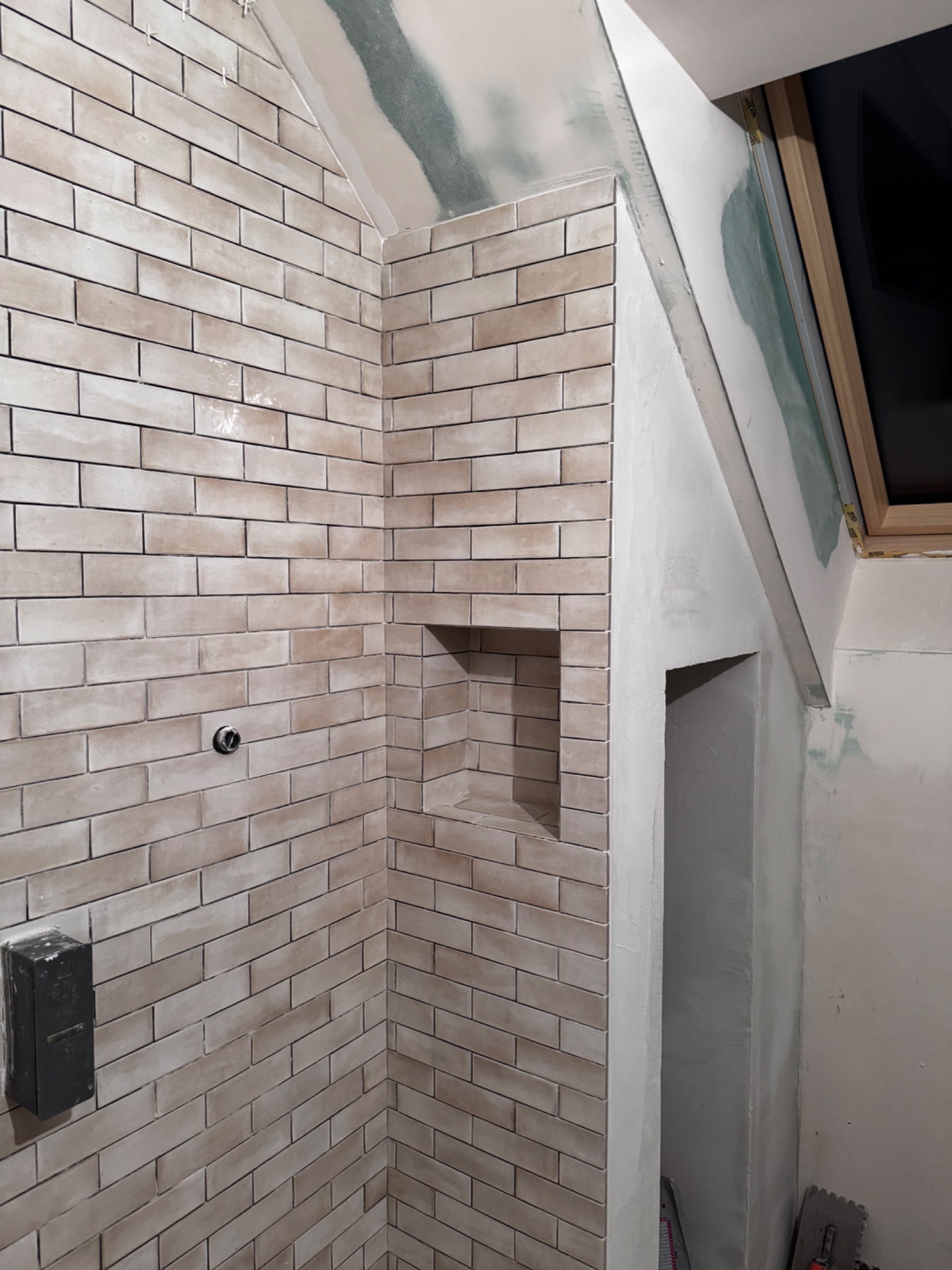 Wykończona ściana z beżowych płytek w łazience na poddaszu, z wnęką i widoczną instalacją. Ściany przygotowane do dalszych prac wykończeniowych.