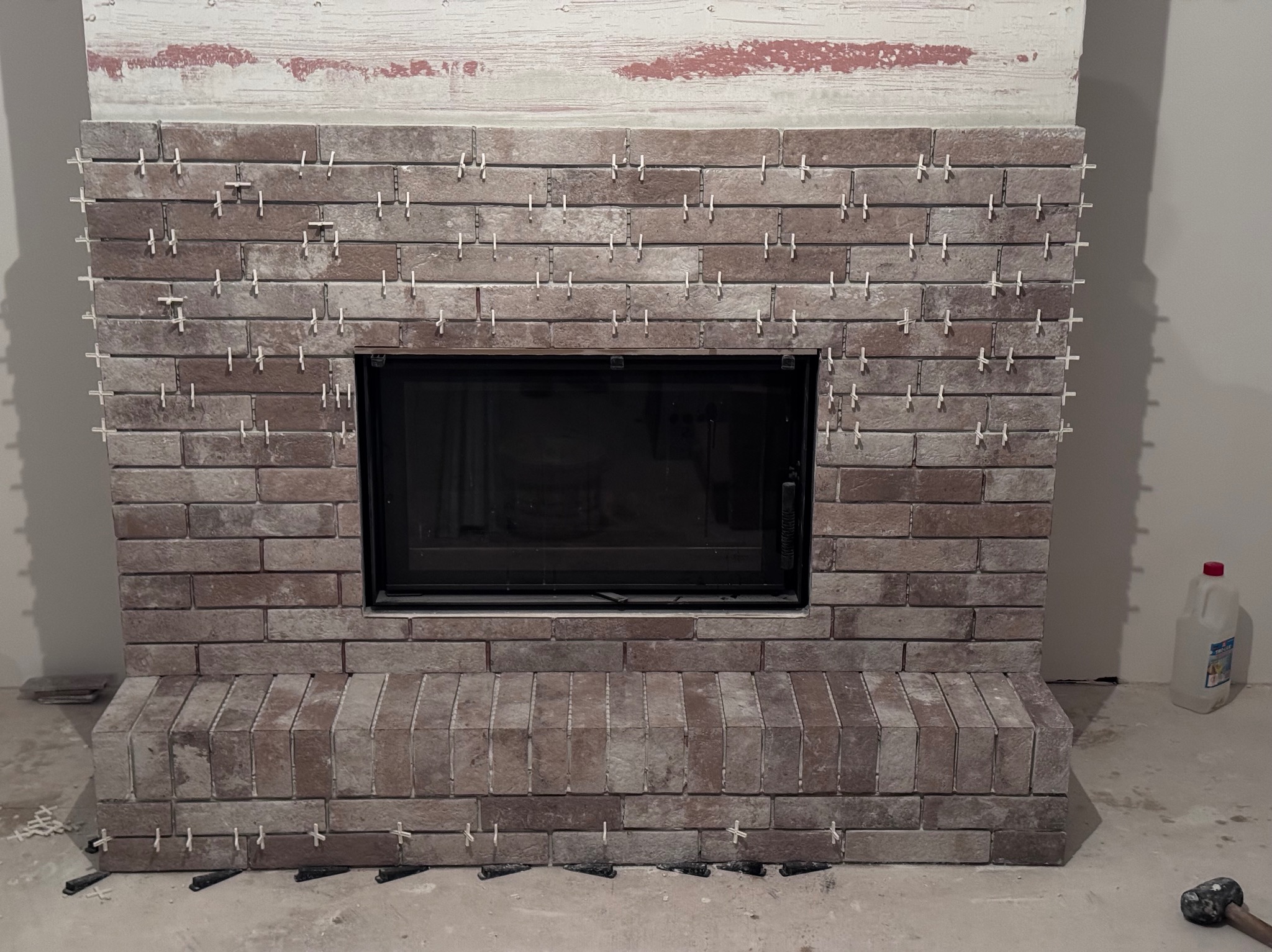 Obudowa kominka z cegieł w trakcie montażu, z krzyżykami dystansowymi. Widoczne narzędzia i materiały budowlane. Ceglana faktura z czarnym wkładem kominkowym.
