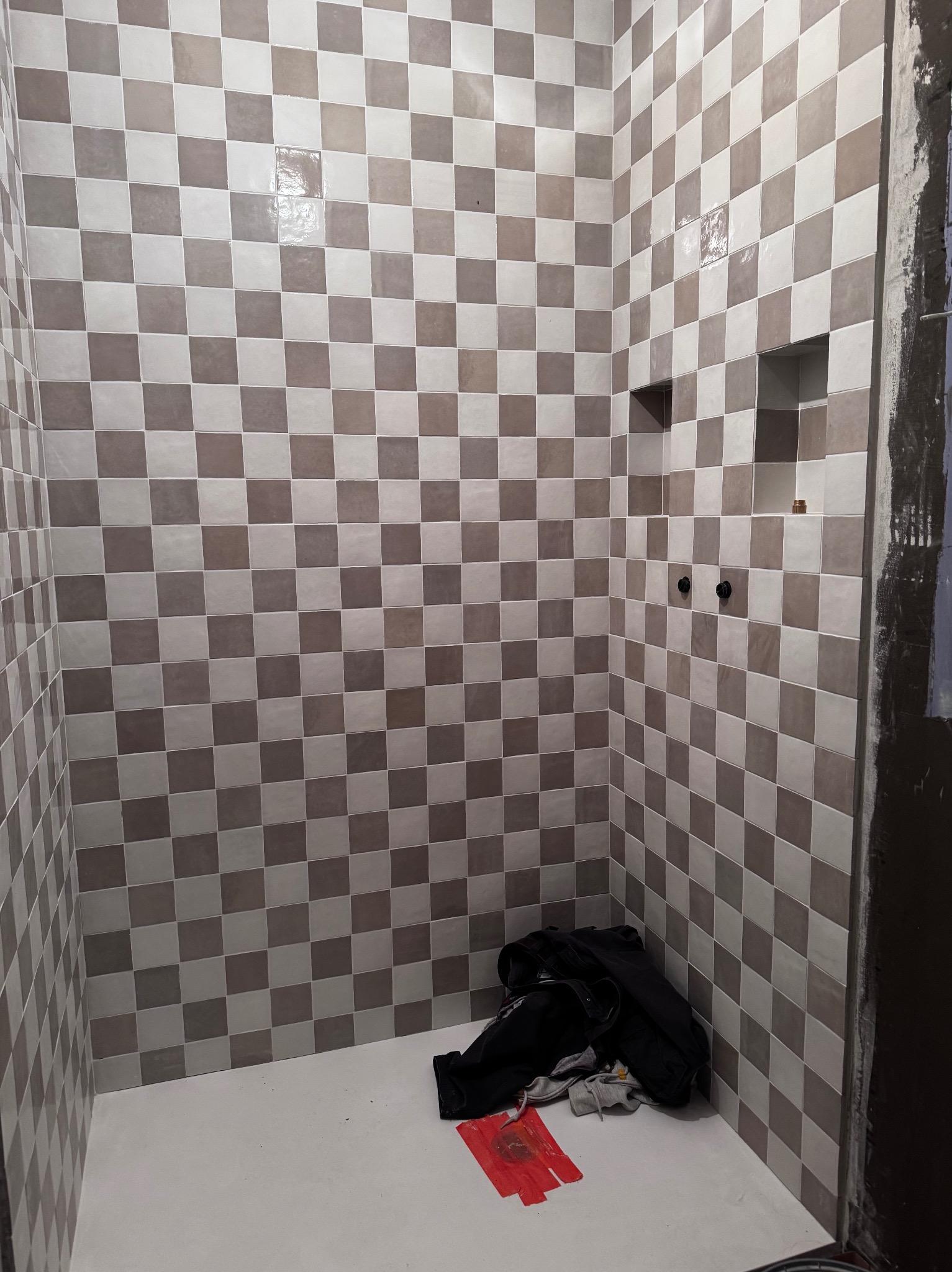 Ściany wyłożone szachownicą z płytek w odcieniach szarości i beżu, z wnęką i wystającymi elementami instalacji. Na podłodze leżą ubrania i narzędzia.