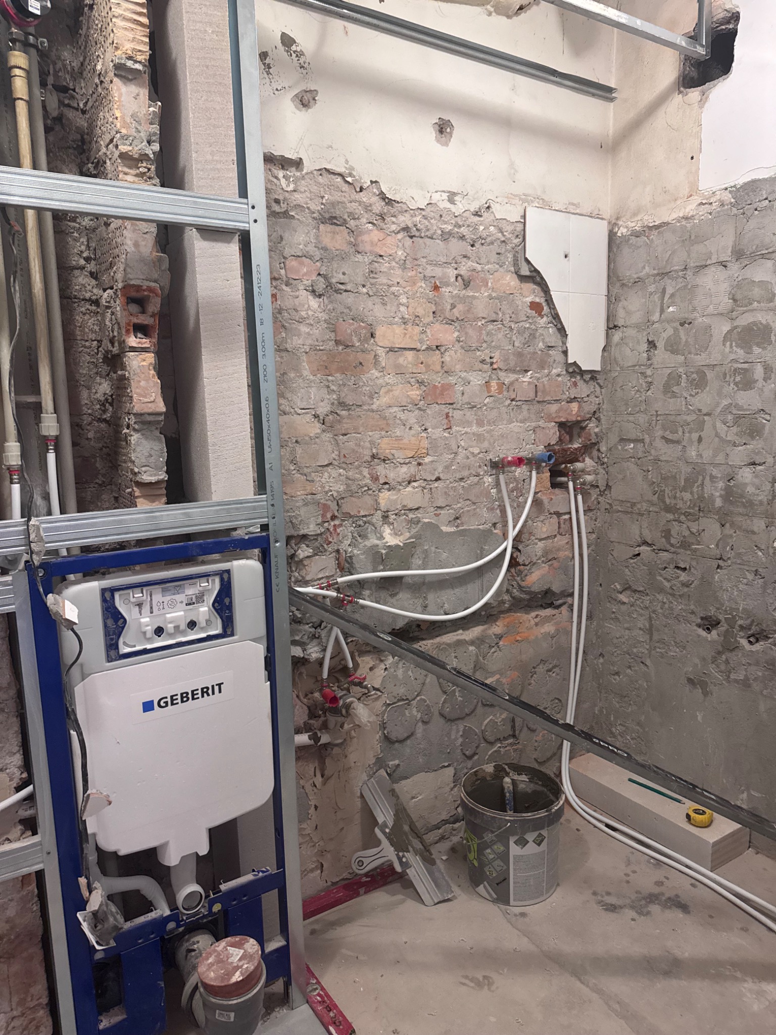 Montaż stelaża podtynkowego Geberit w surowej łazience z odsłoniętą cegłą i instalacją wodną. Widoczne narzędzia i materiały budowlane.