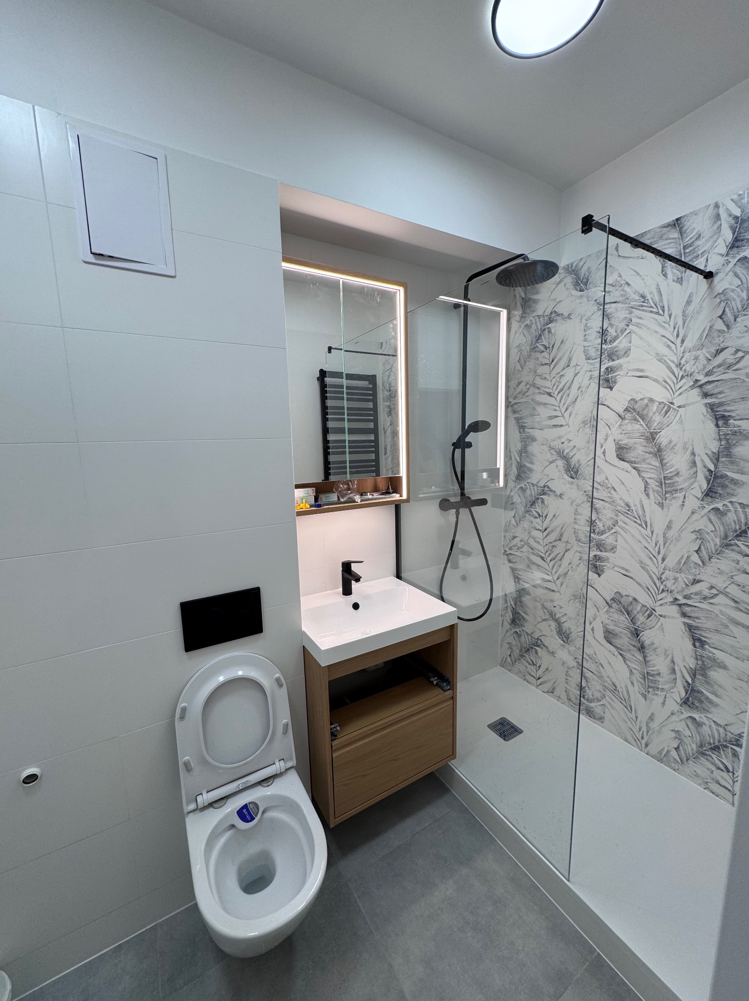 Nowoczesna łazienka po remoncie: białe płytki, kabina prysznicowa z motywem liści, umywalka z szafką z jasnego drewna i podświetlane lustro. Minimalistyczny design.