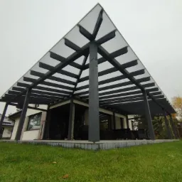 Nowoczesna altana z drewnianą konstrukcją i przezroczystym zadaszeniem, przylegająca do domu z panelami słonecznymi na dachu, widok z poziomu trawnika.