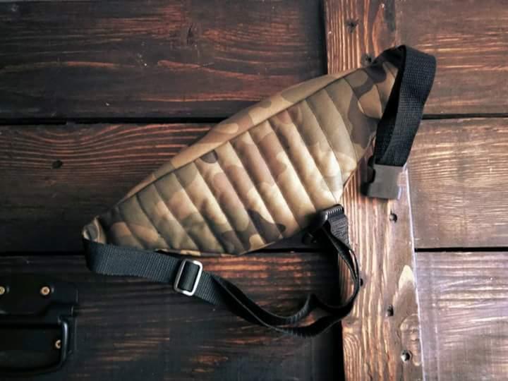 Saszetka nerka z pikowanego materiału w kamuflażu, leżąca na tle ciemnego, drewnianego panelu.