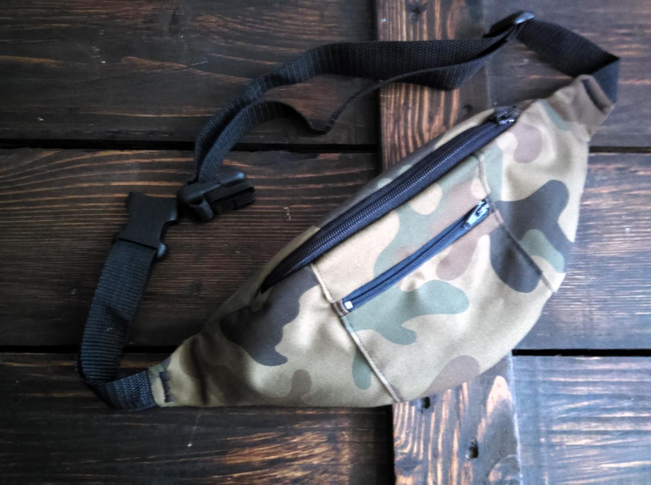 Saszetka biodrowa typu nerka w kamuflażu moro z czarnym paskiem i plastikową klamrą, leżąca na ciemnym, drewnianym tle.