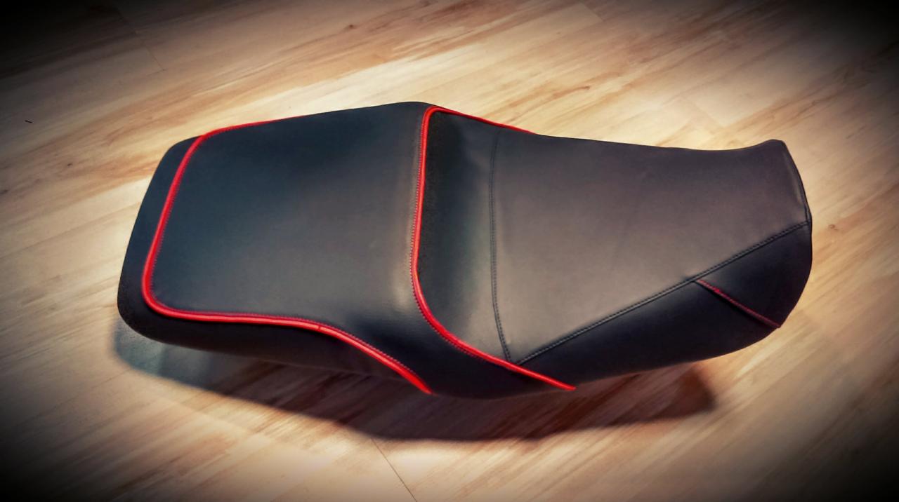 Czarne skórzane siedzisko motocyklowe z czerwonym obszyciem, leżące na drewnianej podłodze, widoczne przeszycia i faktura skóry.