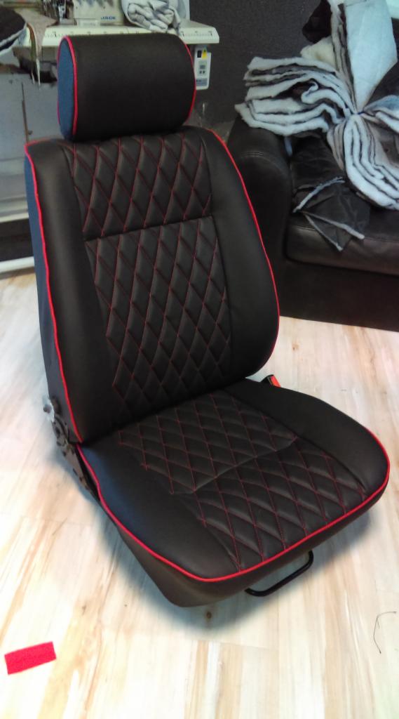 Czarne, pikowane siedzisko samochodowe z czerwoną lamówką, widoczne na tle szwalni z maszyną do szycia i stosem materiałów.