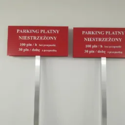 Dwa identyczne czerwone szyldy z białym napisem 'PARKING PŁATNY NIESTRZEŻONY' oraz cennikiem godzinowym i dobowym, zamocowane na srebrnych słupkach na tle jasnej ściany.