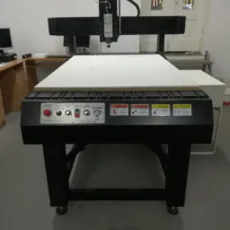 Czarna frezarka CNC z białym arkuszem materiału na stole roboczym, widoczne panele sterowania i ostrzeżenia. W tle biurko i okno.