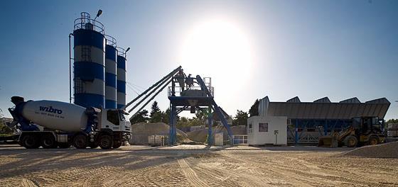 Ciężarówka z betonomieszarką, silosy z niebieskimi pasami, stanowisko załadunkowe i ładowarka na placu budowy betoniarni w słoneczny dzień.