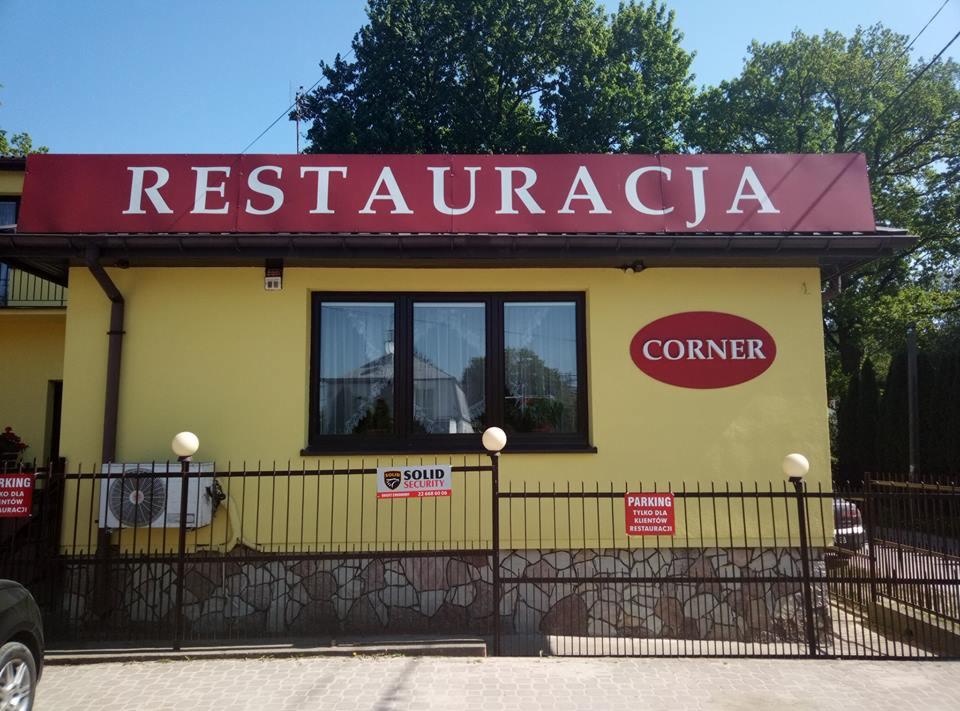 Żółty budynek restauracji z szyldem 'Restauracja' i napisem 'Corner', widoczny parking i ogrodzenie.