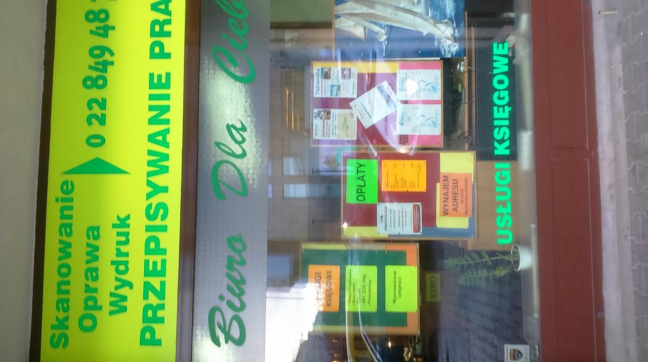 Zewnętrzny widok szyldu biura oferującego usługi skanowania, oprawy, wydruku, przepisywania oraz usługi księgowe, z widocznymi ogłoszeniami na tablicach za szybą.