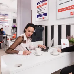 Kobieta w bluzce z siateczkowymi rękawami pisze na dokumentach przy białym biurku z laptopem i filiżankami kawy; w tle widoczne plakaty informacyjne i drukarka.