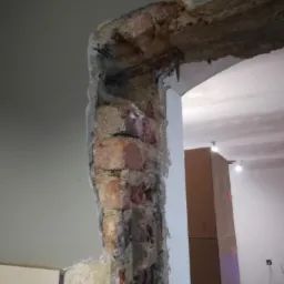 Fragment ściany z odsłoniętą czerwoną cegłą i wystającym zbrojeniem, w trakcie prac przygotowawczych do wykończenia wnętrza.