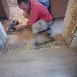 Usługi ogólnobudowlane - Mężczyzna w czerwonej koszulce przycina deskę podłogową za pomocą piły elektrycznej w trakcie prac wykończeniowych, widoczne ślady cięcia, kurz i resztki materiałów.