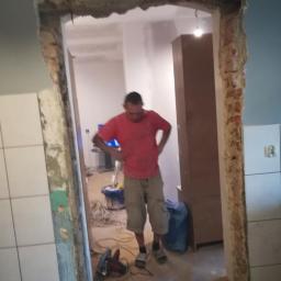 Usługi ogólnobudowlane - Pracownik w czerwonej koszulce stoi w otworze drzwiowym w trakcie remontu, widoczne uszkodzone ściany i narzędzia, w tle wnętrze mieszkania z szafą i telewizorem.