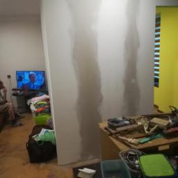 Usługi ogólnobudowlane - Świeżo zagruntowana ściana działowa w trakcie prac przygotowawczych, widoczne ślady po szpachlowaniu, w tle telewizor z włączonym programem oraz porozrzucane przedmioty i narzędzia.