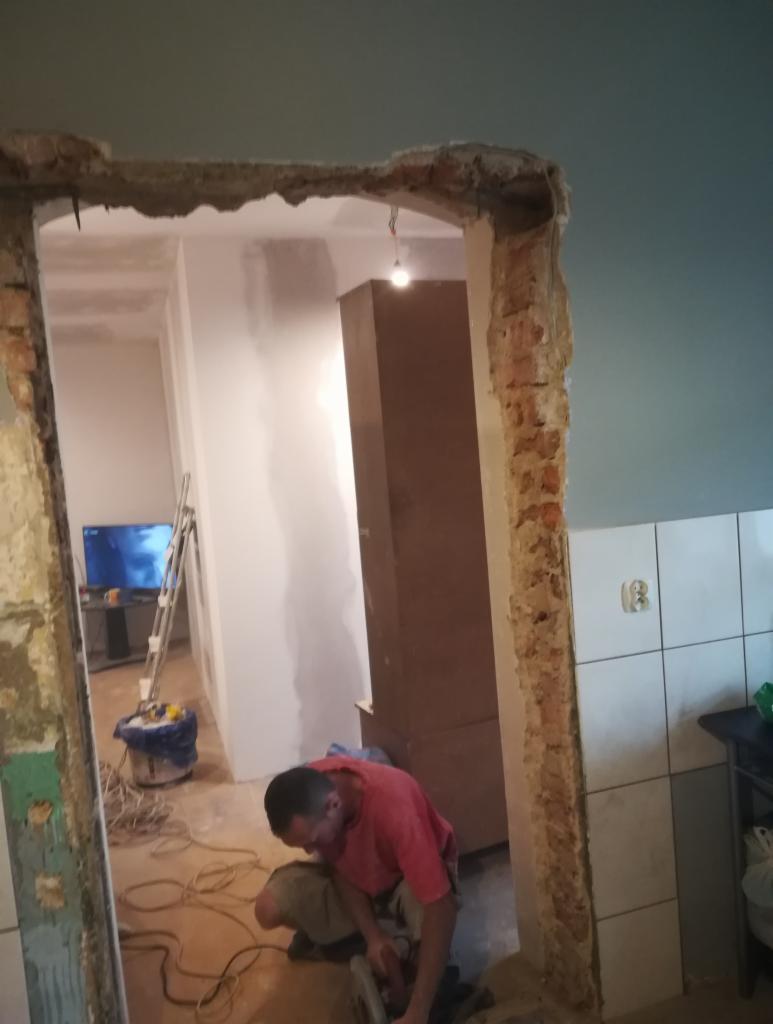 Pracownik w czerwonej koszulce wycina otwór w ścianie z cegły, widoczny remontowany pokój z drabiną i telewizorem w tle.