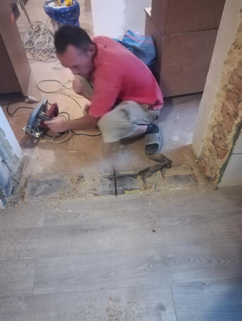 Mężczyzna w czerwonej koszulce przycina deskę podłogową za pomocą piły elektrycznej w trakcie prac wykończeniowych, widoczne ślady cięcia, kurz i resztki materiałów.