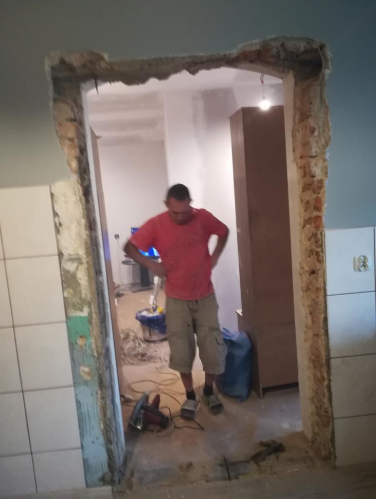 Pracownik w czerwonej koszulce stoi w otworze drzwiowym w trakcie remontu, widoczne uszkodzone ściany i narzędzia, w tle wnętrze mieszkania z szafą i telewizorem.