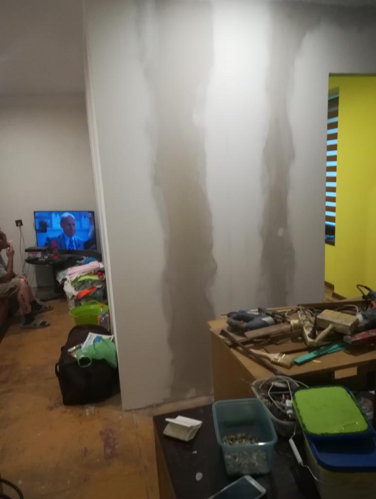 Świeżo zagruntowana ściana działowa w trakcie prac przygotowawczych, widoczne ślady po szpachlowaniu, w tle telewizor z włączonym programem oraz porozrzucane przedmioty i narzędzia.