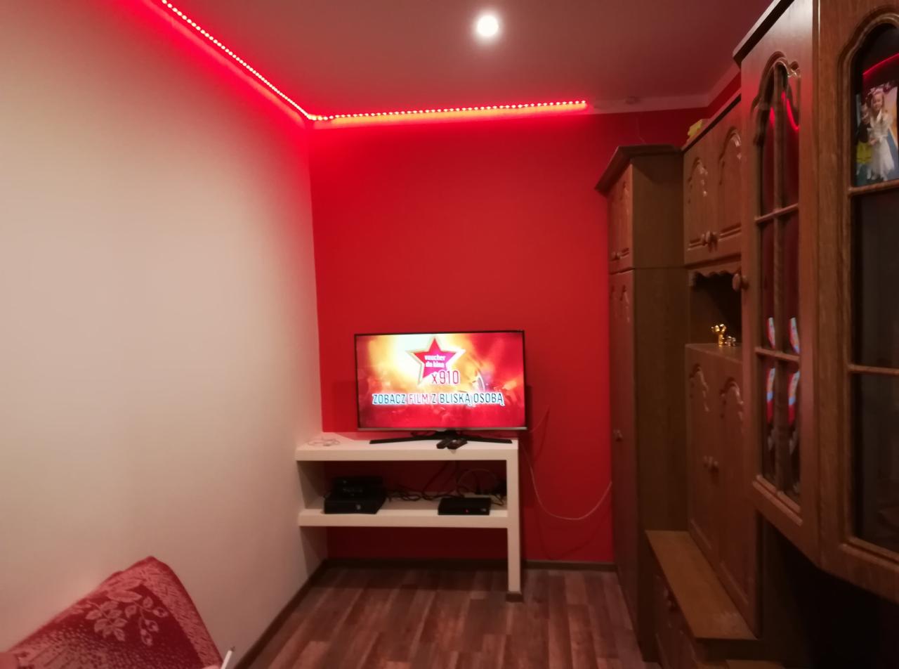 Pokój z czerwoną ścianą, podświetleniem LED na suficie, telewizorem na białej szafce RTV i starą drewnianą szafą.