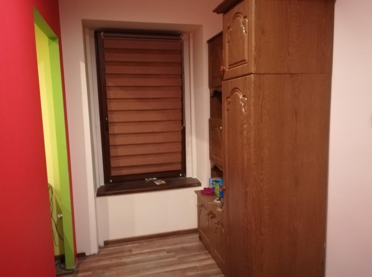 Pokój z brązową drewnianą szafą, poziomymi żaluzjami w oknie, panelami podłogowymi i ścianą w kolorach czerwonym, zielonym i żółtym.