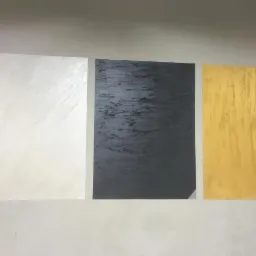 Trzy próbki wykończenia ściany w kolorach: biały z delikatnym połyskiem, grafitowy z wyraźną fakturą i złoty z pionowymi smugami, prezentowane na jasnej ścianie.