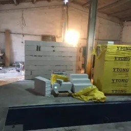 Wnętrze w trakcie budowy: ściana z bloczków Ytong częściowo wzniesiona, paleta z zapakowanymi bloczkami Ytong w żółtej folii, rozpakowane bloczki Kreisel na palecie, surowy beton na podłodze...