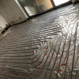 Montaż ogrzewania podłogowego: rury grzewcze ułożone na folii z siatką w surowym wnętrzu, widoczna drabina malarska przy ścianie.