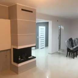 Wnętrze salonu po remoncie: szara ściana, zabudowany kominek z czarnym wkładem, biała podłoga z płytek, krzesła przykryte folią ochronną.