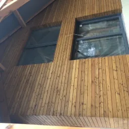 Fragment elewacji budynku z drewnianą okładziną pionową i dwoma oknami, widoczne elementy konstrukcji dachu i ocieplenie styropianem po prawej stronie.