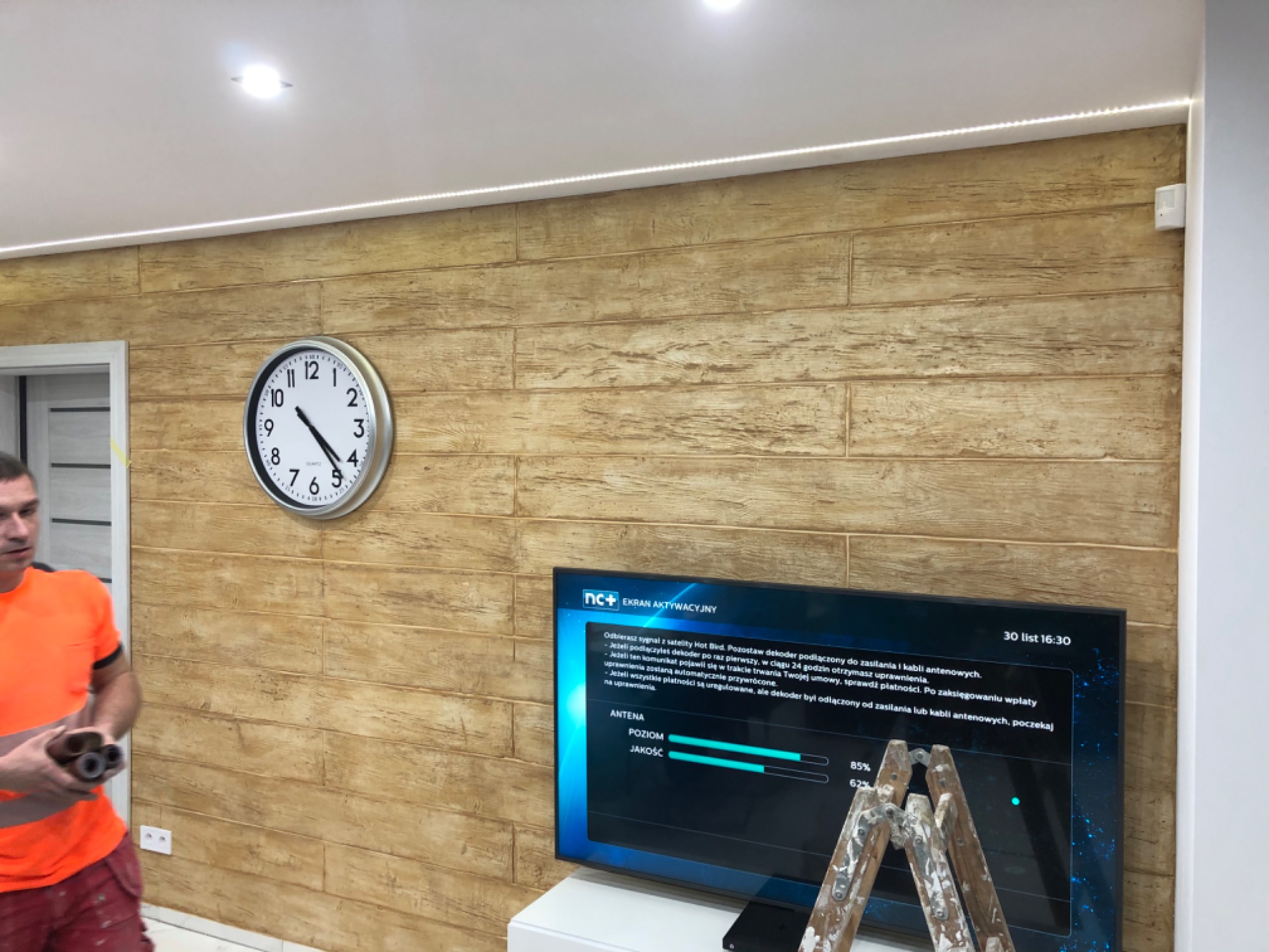 Ściana wykończona panelami imitującymi drewno, z zegarem na ścianie, telewizorem i drabiną malarską w pokoju, widoczna listwa LED pod sufitem oraz fragment sylwetki mężczyzny trzymającego rolki