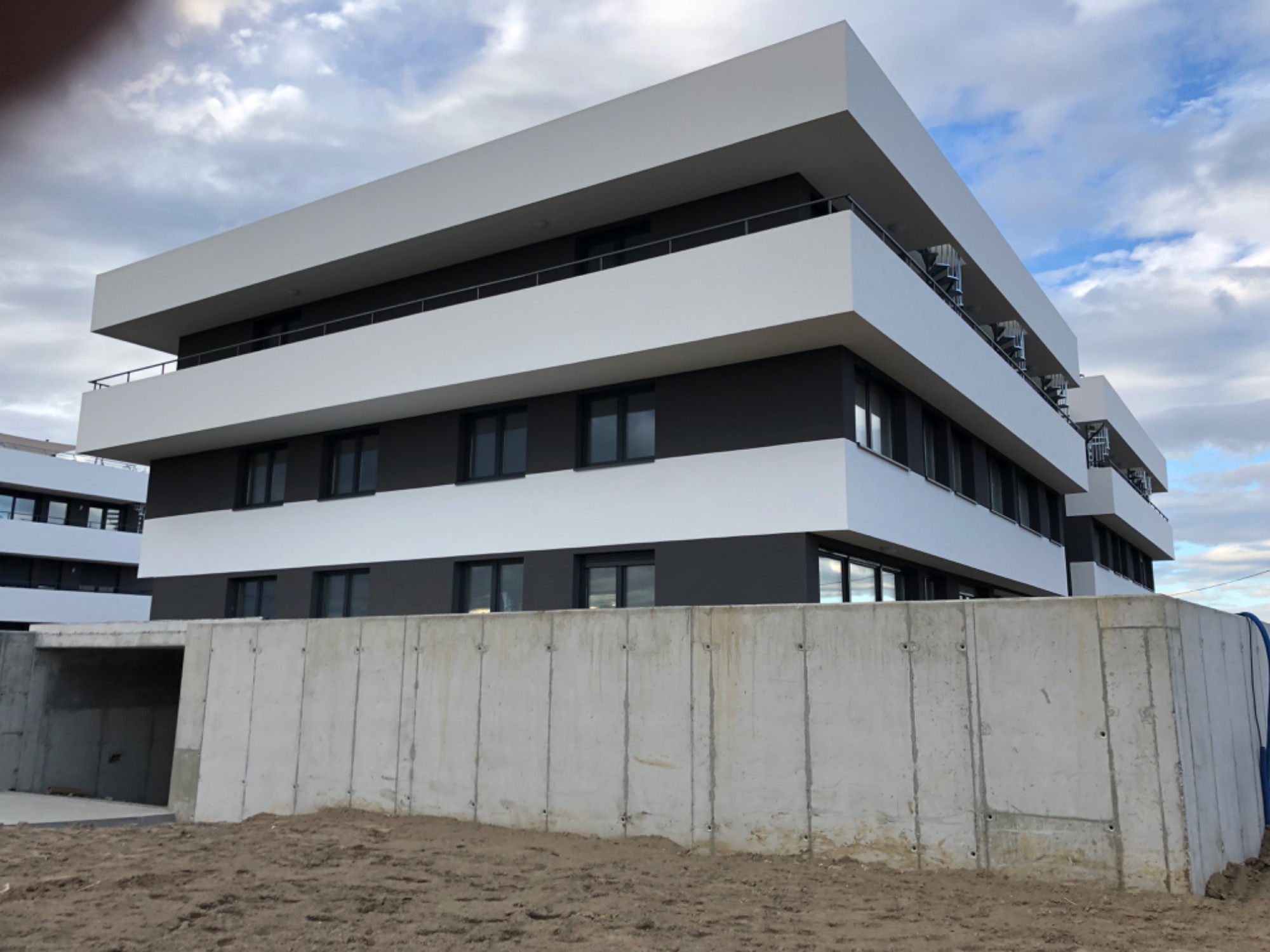 Nowoczesny budynek wielorodzinny z szarą elewacją i białymi elementami architektonicznymi, widoczny z perspektywy ziemi, z surowym betonowym murem w pierwszym planie i pochmurnym niebem w tle.