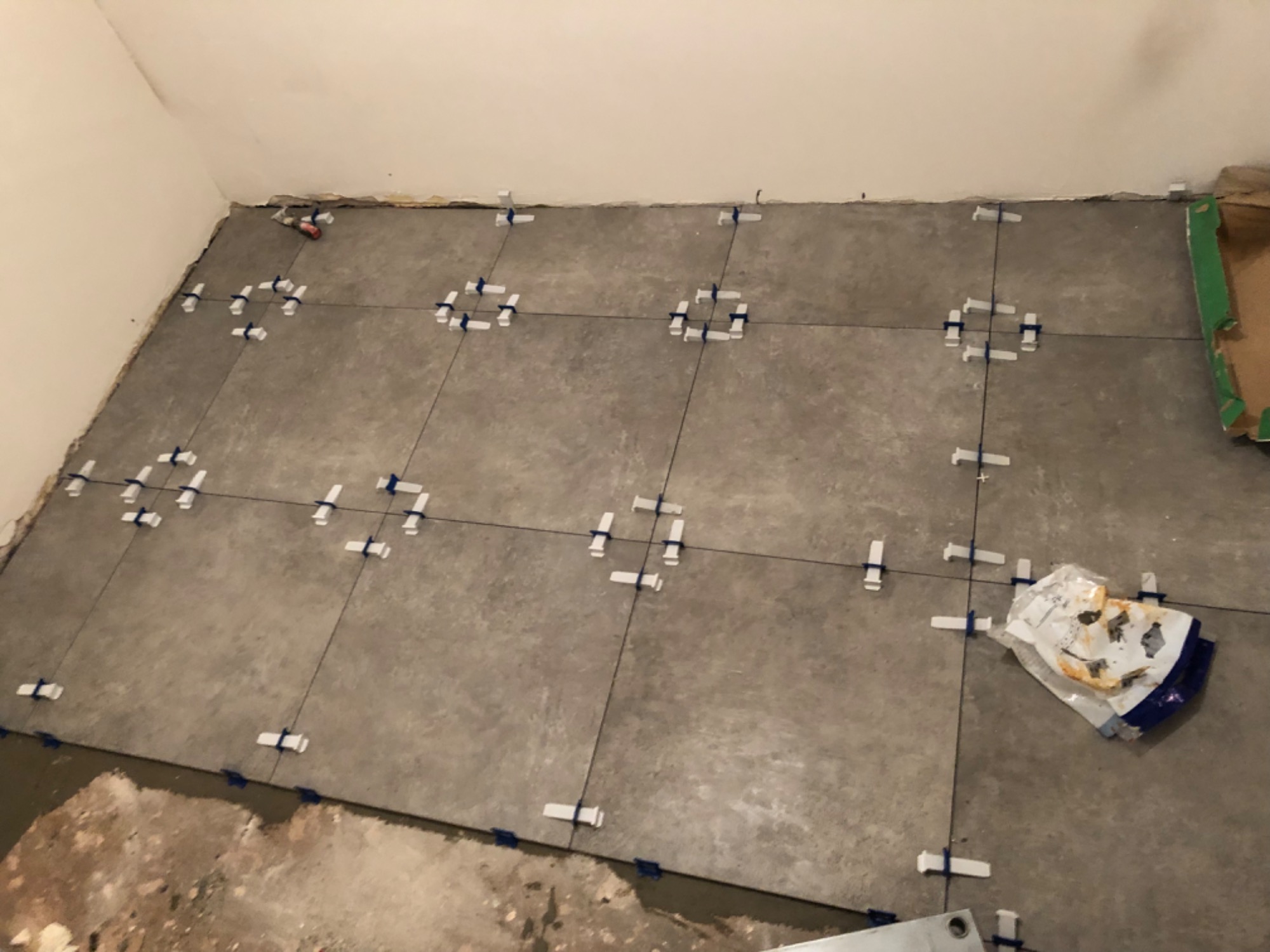 Szare płytki podłogowe w trakcie układania, krzyżyki dystansowe w spoinach, widoczny fragment podłoża przed wykończeniem i zużyte opakowanie po kleju.
