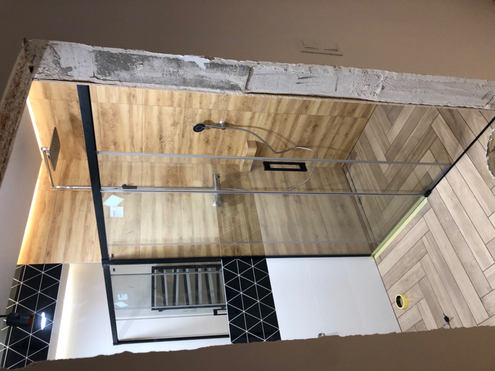 Niedokończona łazienka z drewnopodobnymi płytkami na ścianach i podłodze ułożonymi w jodełkę, częściowo zamontowana szklana kabina prysznicowa z czarnymi profilami, widoczne niedoskonałości...