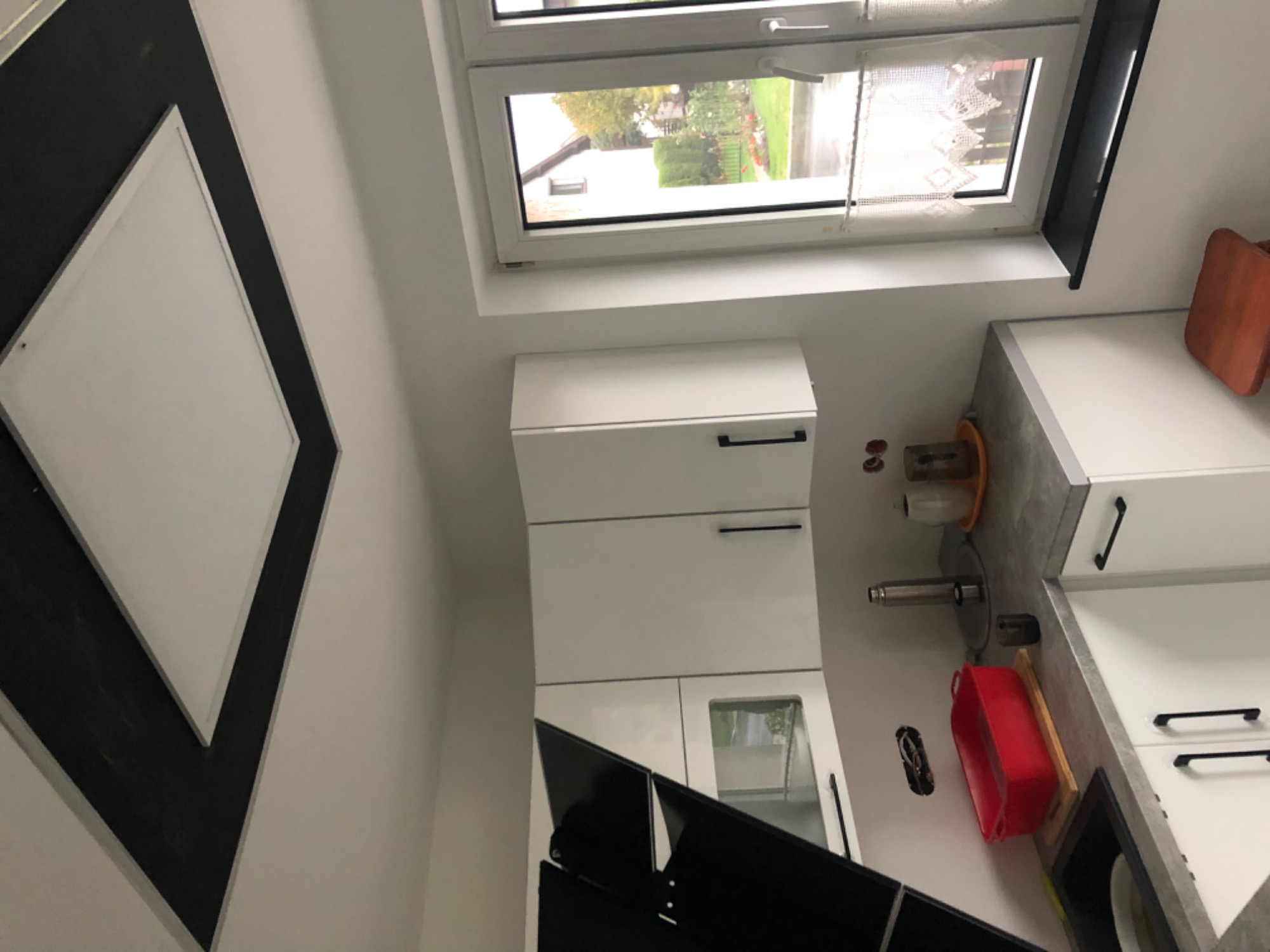 Nowoczesna, biała kuchnia z szarym blatem i czarnymi uchwytami, widziana z nietypowej perspektywy, okno z widokiem na zieleń w tle, dekoracyjny element na ścianie w czarno-białej kolorystyce.