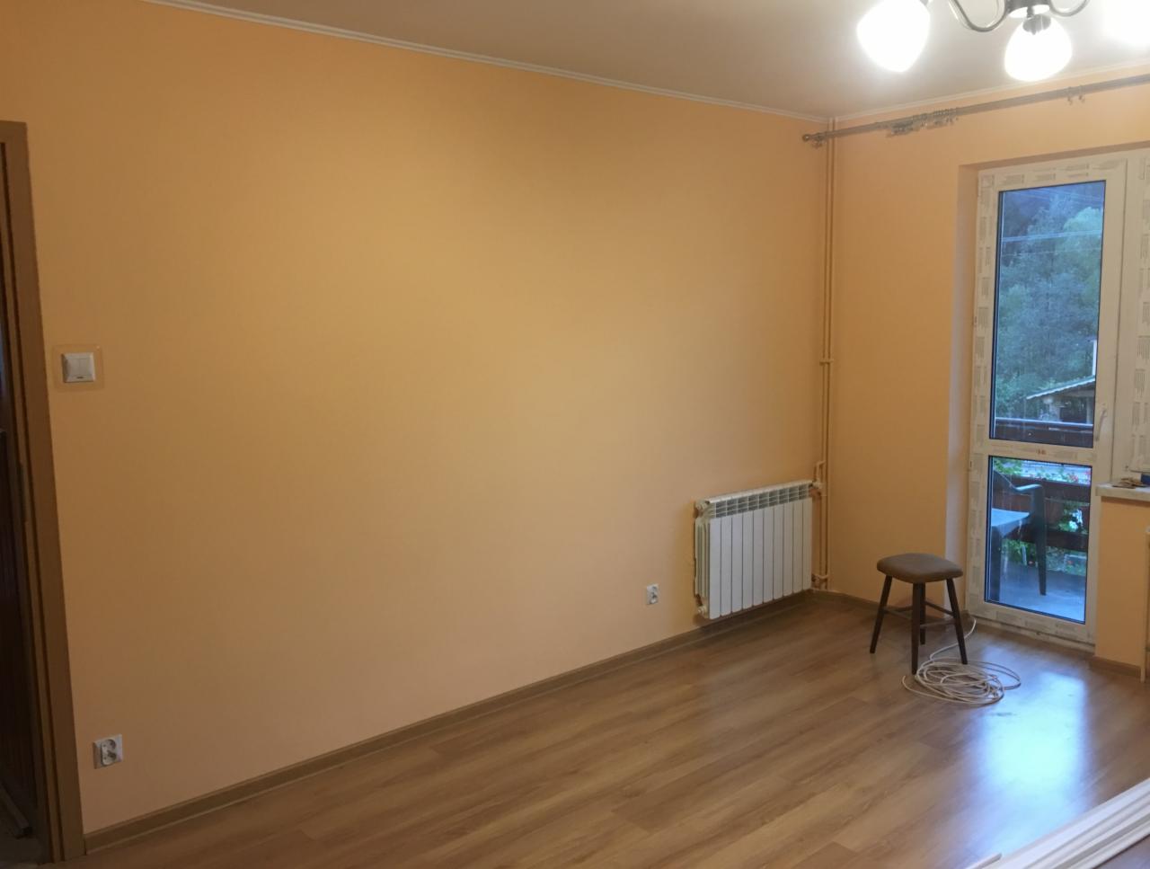 Pokój po malowaniu ścian na kolor brzoskwiniowy, z drewnianą podłogą, białym grzejnikiem, krzesłem i zwiniętym kablem elektrycznym na podłodze.