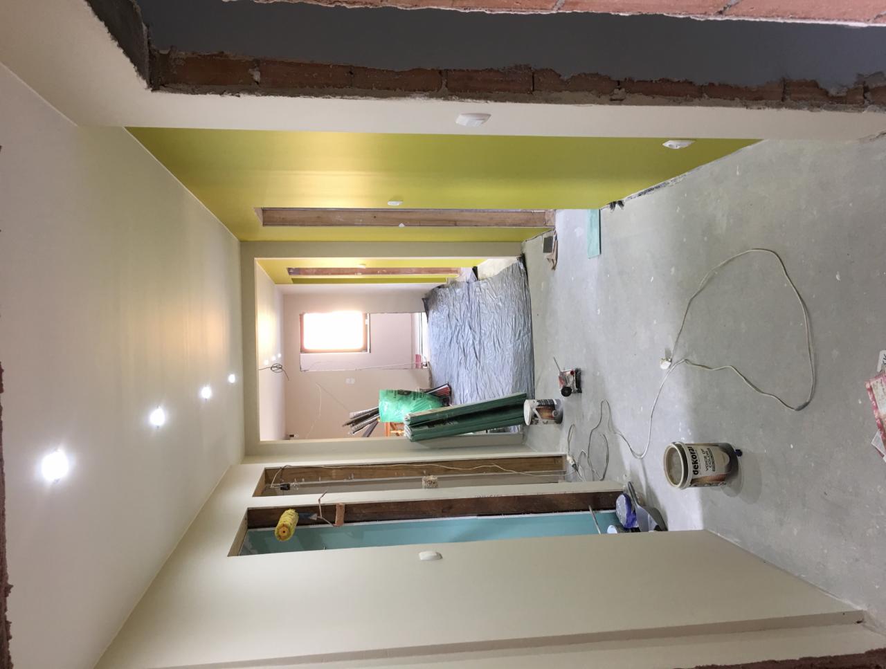 Korytarz w trakcie prac wykończeniowych: pomalowane ściany w odcieniach kremu i limonki, surowy beton na suficie, widoczne puszki z farbą Dekoral i narzędzia malarskie na podłodze zabezpieczonej...