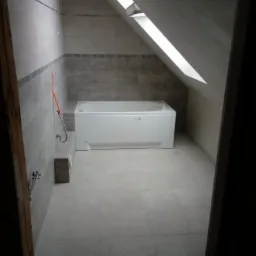 Łazienka na poddaszu z oknem dachowym, wykończona szarymi płytkami, z białą wanną.