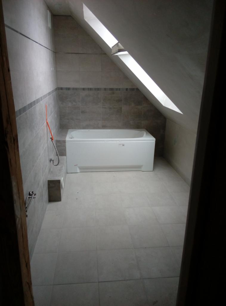 Łazienka na poddaszu z oknem dachowym, wykończona szarymi płytkami, z białą wanną.