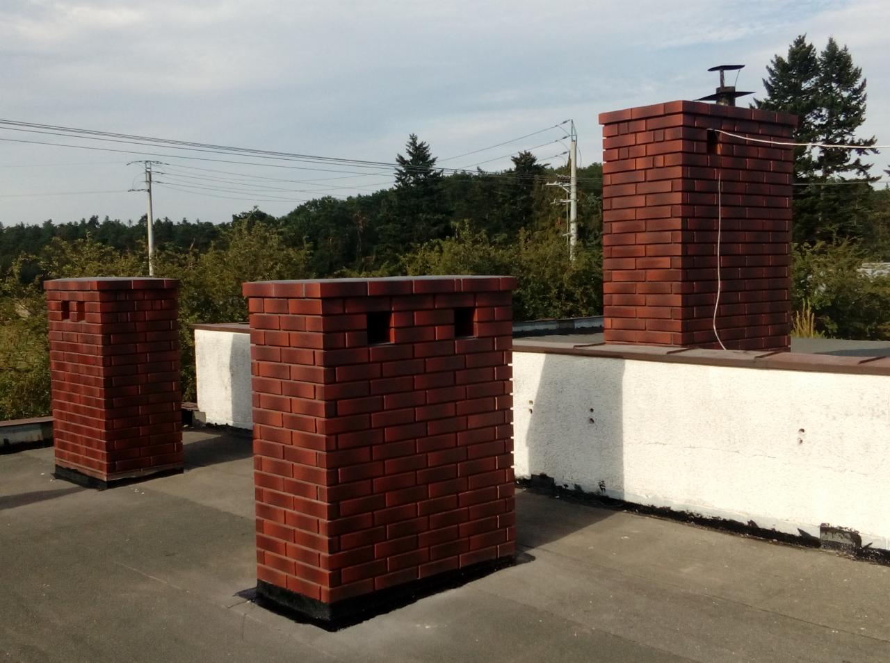 Trzy kominy z czerwonej cegły na płaskim dachu pokrytym czarną papą, na tle zieleni drzew i pochmurnego nieba.