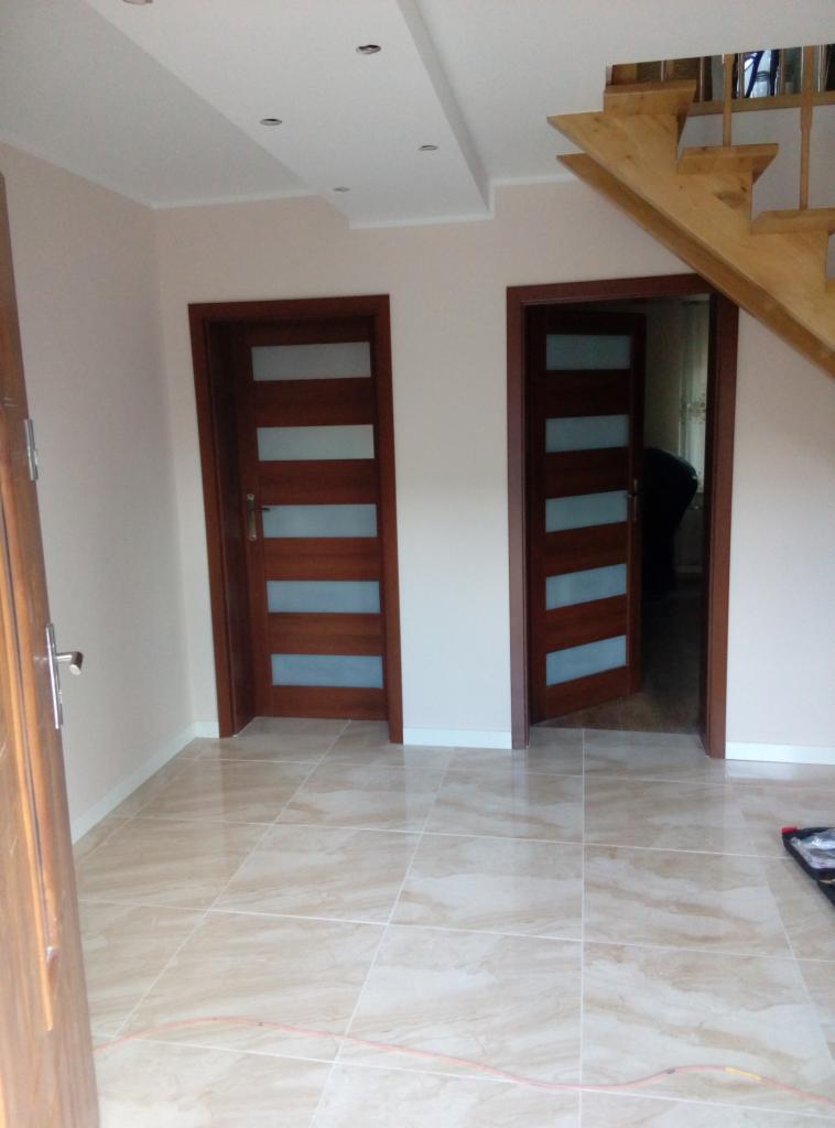 Jasne wnętrze po remoncie z beżową podłogą z płytek, dwiema drewnianymi drzwiami z przeszkleniami i fragmentem drewnianych schodów.