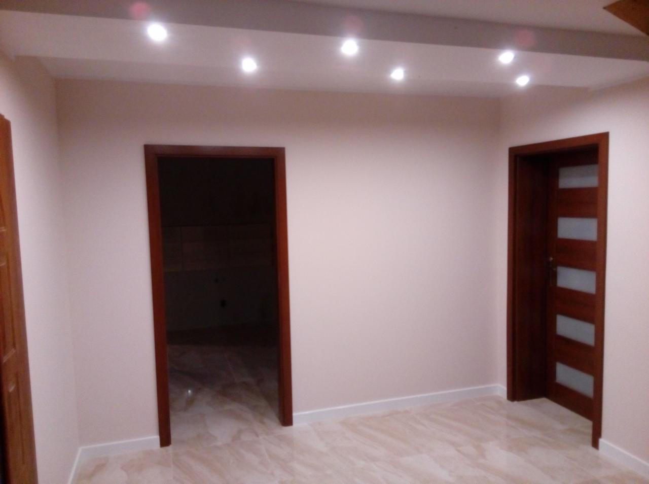 Pomalowane na jasny kolor wnętrze z dwiema ciemnymi drewnianymi ościeżnicami; jedna z pełnymi drzwiami z przeszkleniami, druga otwarta na sąsiednie pomieszczenie, marmurowe płytki podłogowe...